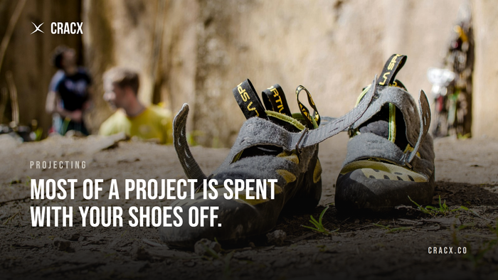 Worn climbing shoes left on the dirt at the base of a crag while climbers rest in the background