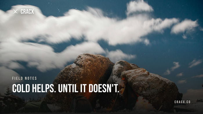 Granite boulders dusted with snow under a night sky, a small campfire glowing at their base and two tents beside them