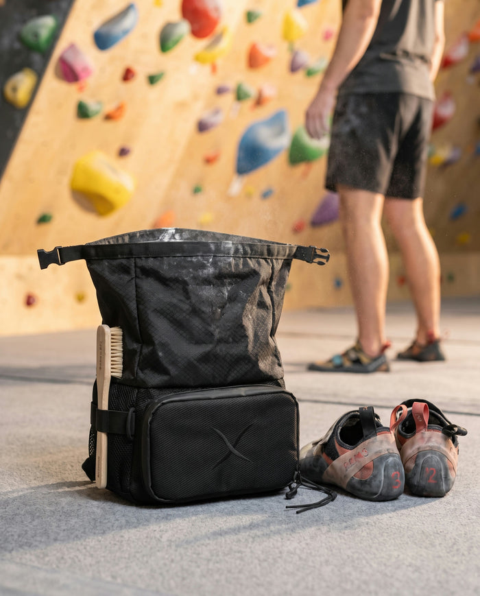The CRACX Ascend open on a bouldering gym floor next to a pair of rental climbing shoes, a climber at the wall behind