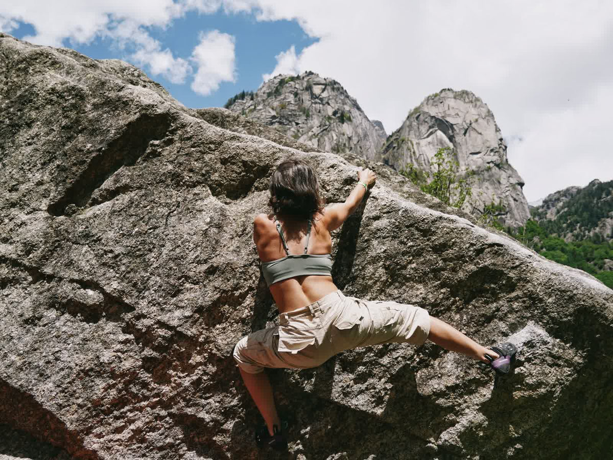 A boulderer topping out a granite boulder with a mountain cirque behind her
