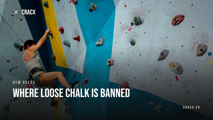 A boulderer with chalked hands mid-move on an indoor gym wall, chalk bag on her waist