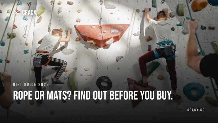 Two young climbers on a top-rope wall at an indoor climbing gym, each wearing a harness with a chalk bag clipped on, while belayers hold the ropes