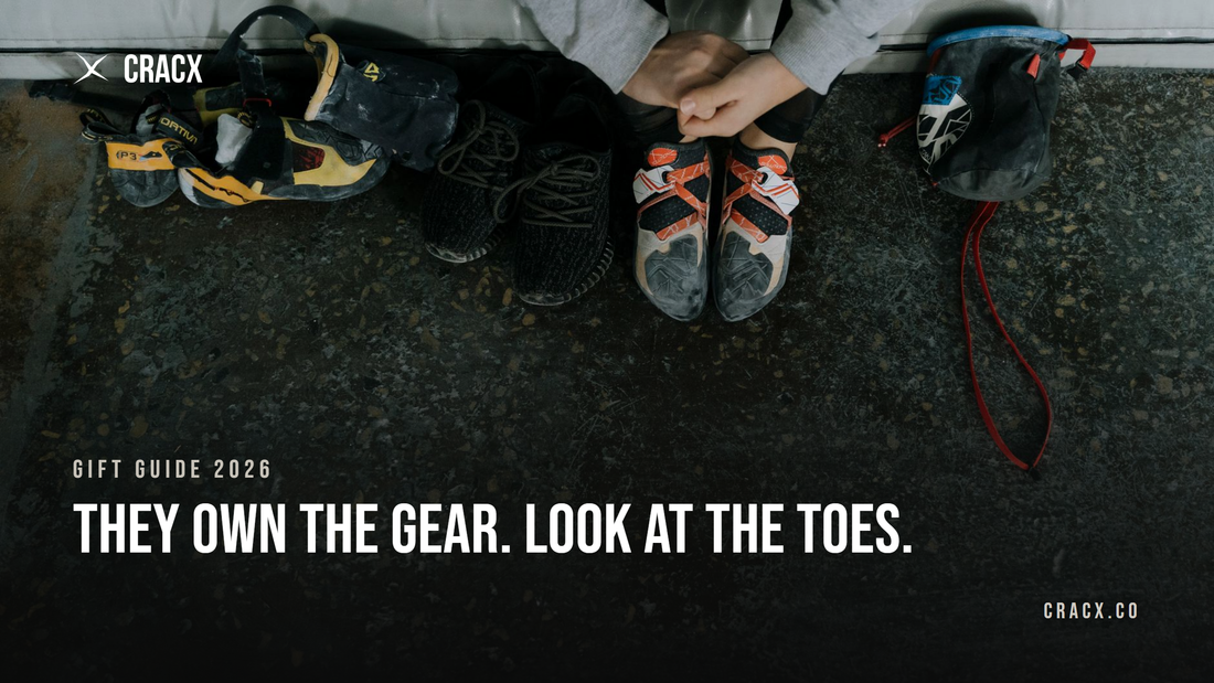 A climber sitting on a gym crash pad wearing worn climbing shoes, with a second pair of climbing shoes, sneakers and a chalk bag on the concrete floor