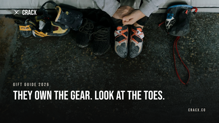 A climber sitting on a gym crash pad wearing worn climbing shoes, with a second pair of climbing shoes, sneakers and a chalk bag on the concrete floor