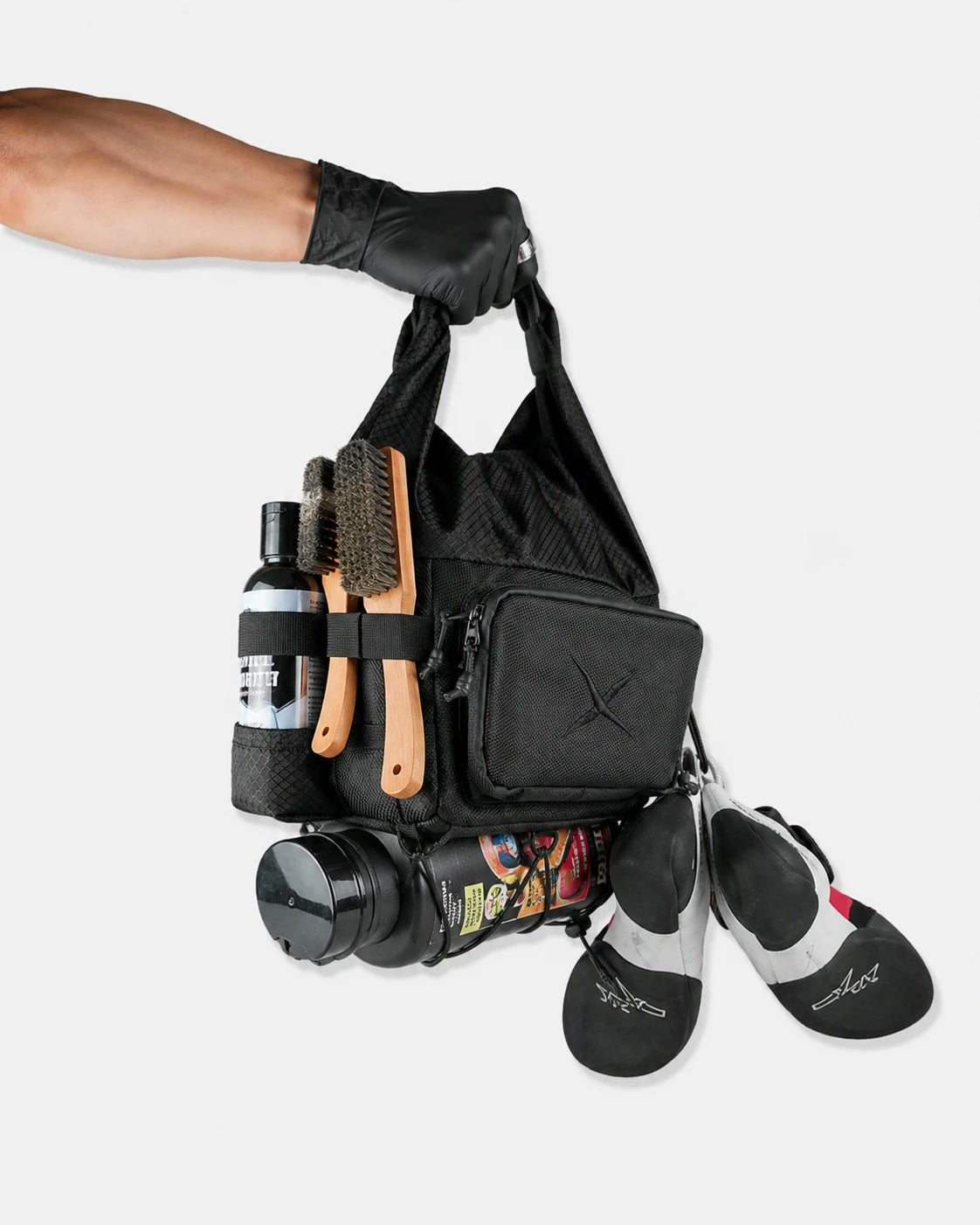 CRACX Ascend bouldering chalk bucket carried by its roll-top handle, fully loaded: two brushes and a liquid chalk bottle on the side, shoes and a water bottle held under the bungee cord
